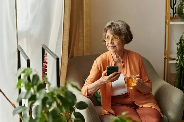 Mujer mayor sonríe mientras disfruta del té y el uso de su smartphone en una acogedora sala de estar. - foto de stock