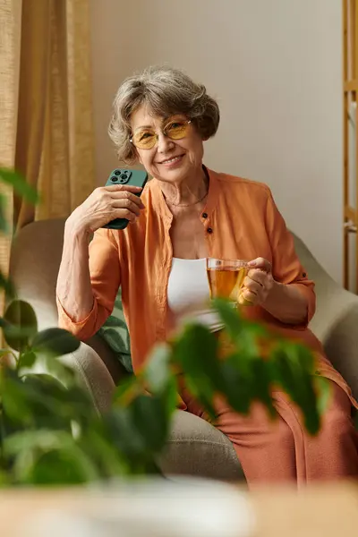 A mature woman relaxes on her comfortable couch, holding a warm drink and smiling at her phone. — Stock Photo