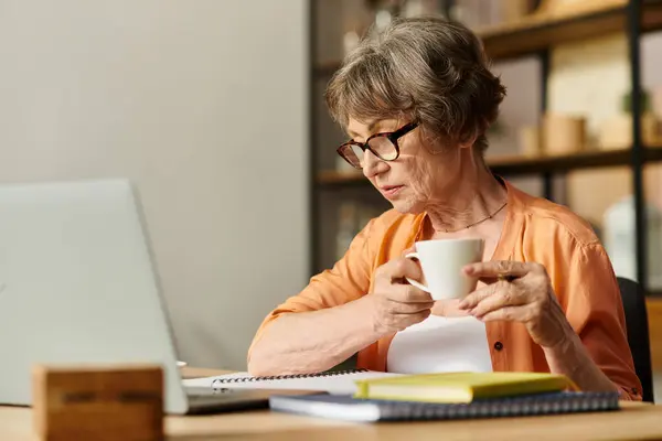 Mature woman relaxes in cozy attire while sipping tea and working on her laptop at home. — Stock Photo