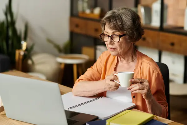 Mature woman in comfortable attire sits at a table, sipping tea while focused on her laptop. — Stock Photo