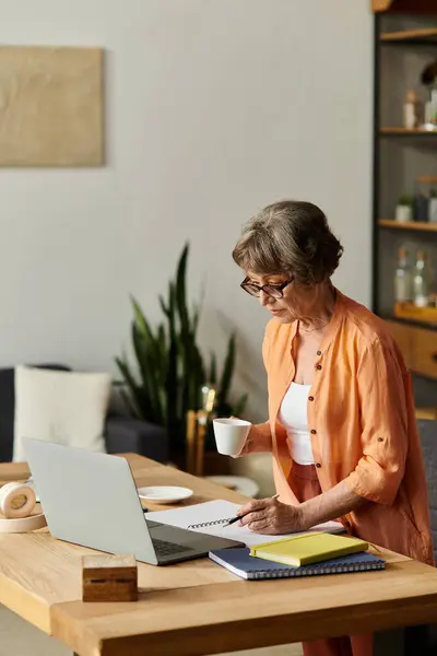 A senior woman dressed in cozy attire works on her projects at home, sipping a warm drink. — Stock Photo