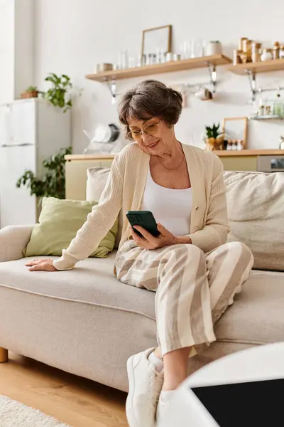 Senior woman in comfy clothes smiles on sofa, using her smartphone in a cozy setting. — Stock Photo