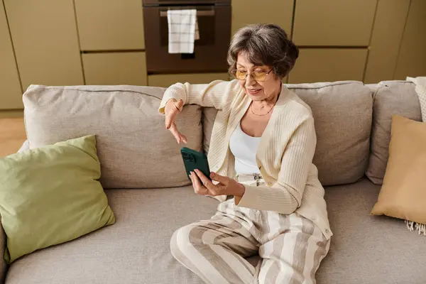 Mature woman relaxes on her sofa, scrolling through her smartphone in a cozy living space. — Stock Photo