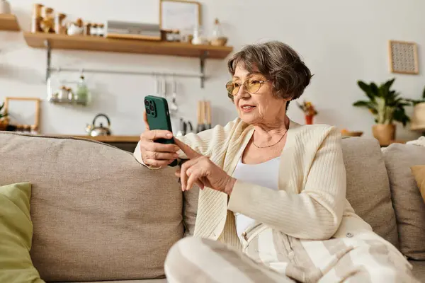 Mature woman wearing cozy attire relaxes on a couch, engaging with her smartphone in comfort. — Stock Photo