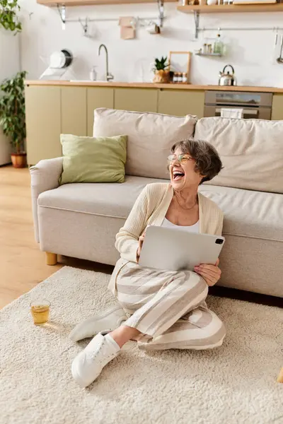 A senior woman in comfy attire laughs with her tablet while sitting on the floor. — Stock Photo
