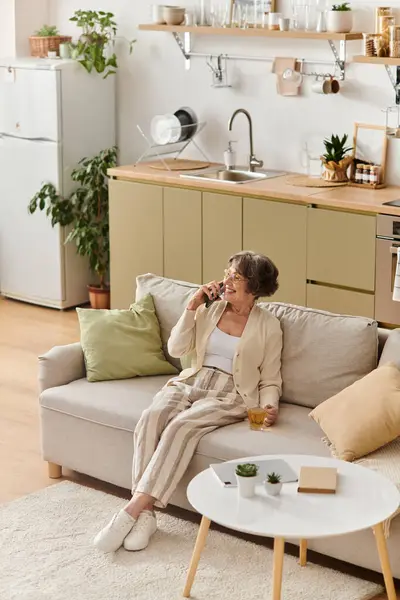 A senior woman comfortably sits on a sofa, sipping a drink and enjoying her serene home environment. — Stock Photo