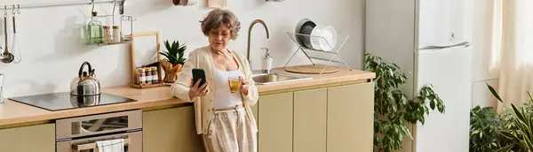 A senior woman in cozy attire relaxes in her beautifully arranged kitchen, sipping a drink. — Stock Photo