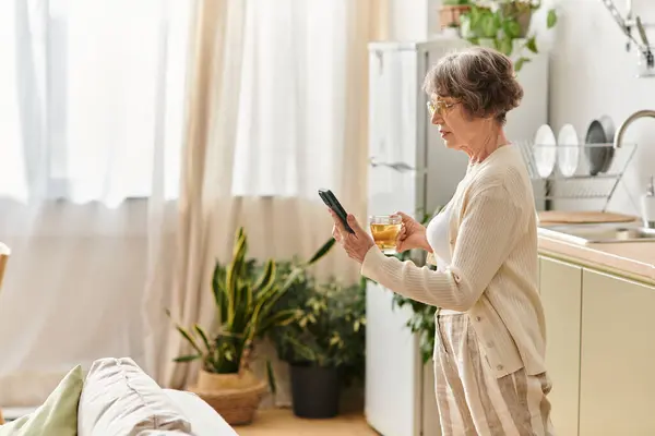 Una mujer mayor en ropa acogedora disfruta de una bebida caliente en casa mientras usa su teléfono. - foto de stock