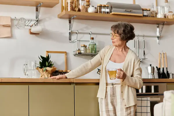 Senior woman in cozy attire stands in her kitchen while savoring a warm cup of tea. — стоковое фото