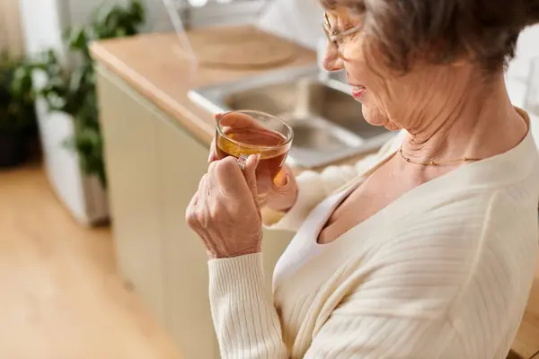 A mature woman in cozy attire enjoys her warm drink in the comfort of her kitchen. — Stock Photo