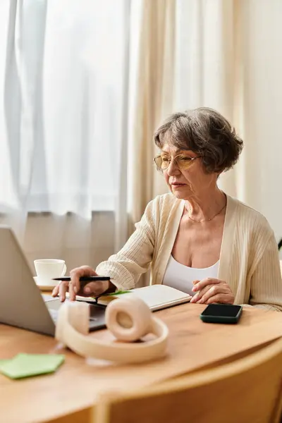 A mature woman relaxes at her home desk, engaged with her laptop and notes while being cozy. — Stock Photo