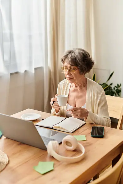 Mature woman in comfy attire savors tea as she engages with her laptop in a cozy setting. — Stock Photo