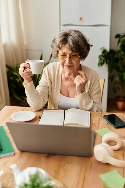 Mature woman relaxes in a comfortable setting, sipping tea while engaging with her laptop. — Stock Photo