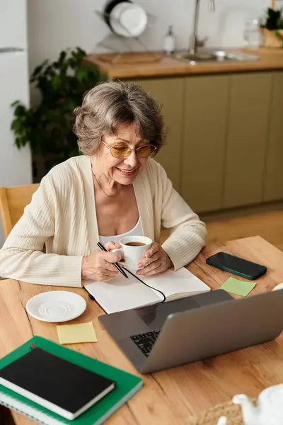 A joyful senior woman sits at a wooden table, sipping coffee and focusing on her laptop with notes. — Stock Photo