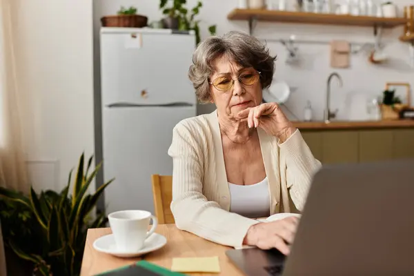A senior woman in cozy clothes sits at her kitchen table, focused on her laptop. — Stock Photo