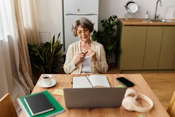 Senior woman in cozy attire sits at a wooden table, engaging with her laptop while enjoying tea. — Stock Photo