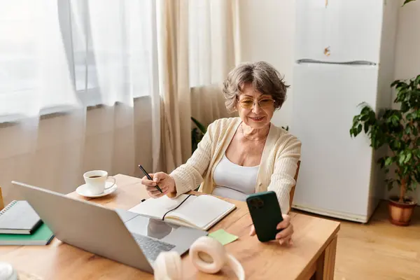 A senior woman relaxes at her desk, engaged with her smartphone and enjoying coffee. — Stock Photo