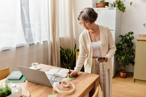 A senior woman in cozy clothes takes notes at a wooden table with her laptop. — Stock Photo
