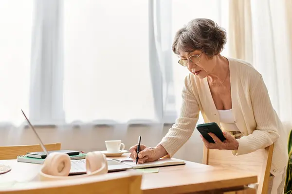 A senior woman enjoys a cozy afternoon at home, jotting down thoughts while holding her phone. — Stock Photo