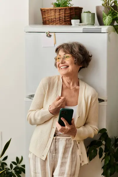 Senior woman in cozy attire stands by her refrigerator, smiling as she uses her smartphone. — Stock Photo