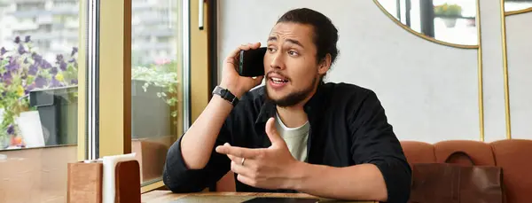 Young man with a beard shares ideas over the phone while working in a lively cafe environment, banner — стоковое фото