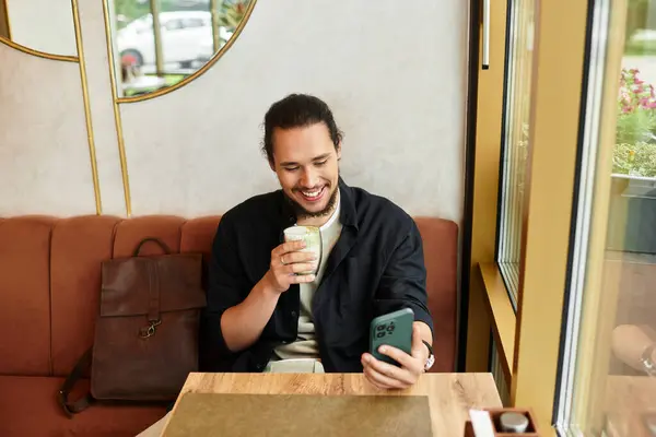 Joven sonríe mientras disfruta de una bebida matcha y el uso de teléfono inteligente en un café soleado. - foto de stock