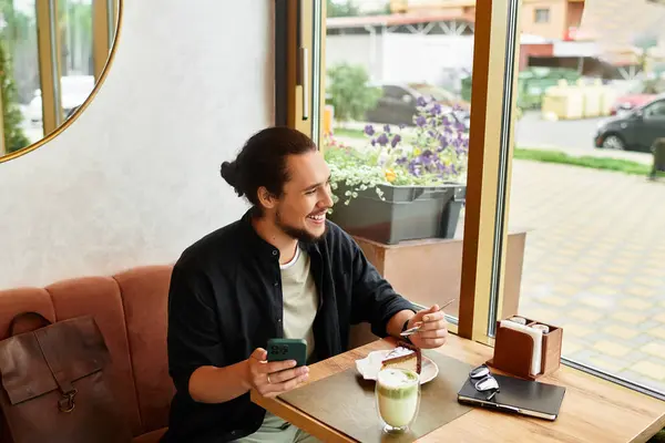 A man with a beard smiles while using smartphone at a cafe, enjoying matcha and cake. — Stock Photo