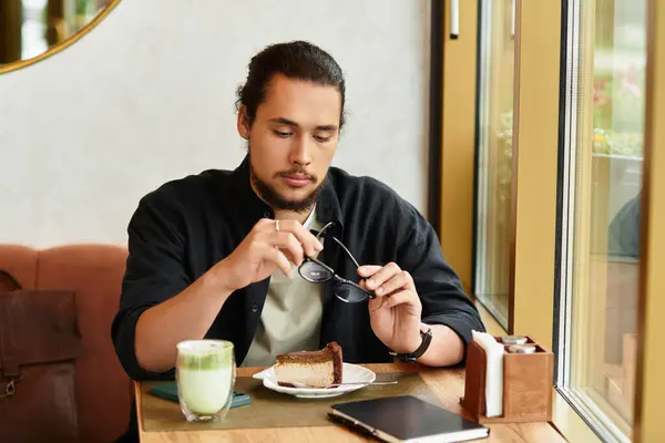 A young man with a beard sits at a cafe table with matcha and cake. — Stock Photo