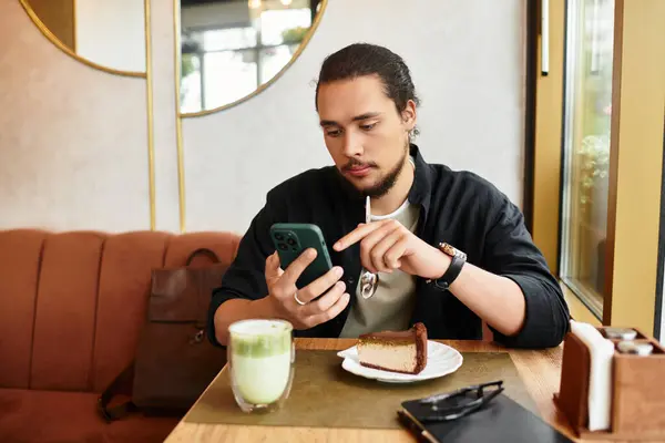 Young man with a beard works on his phone at a cafe, enjoying a matcha and slice of cake. — Stock Photo