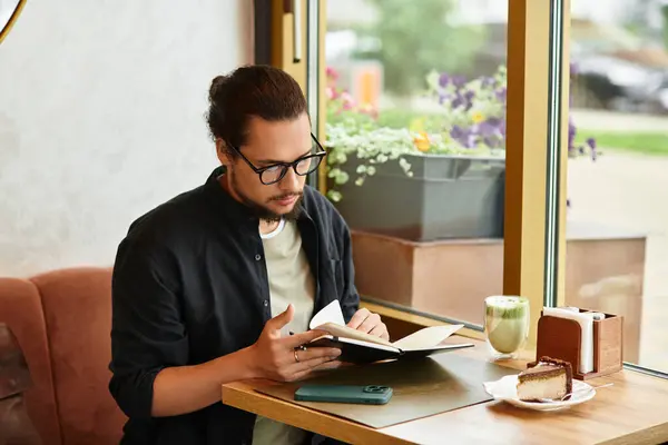 Young man with a beard focuses on his work while sipping matcha and indulging in cake. — Stock Photo