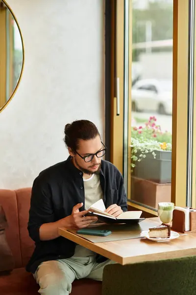 A young man with a beard focuses on his work while sipping matcha and savoring cake in a cafe. — Stock Photo