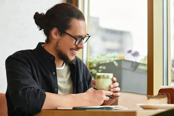A young man with a beard works happily in a cafe, sipping matcha and enjoying cake during summer. — Stock Photo