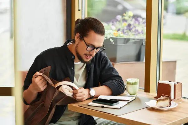 A young man with a beard sits in a cafe, holding bag with headphones — Stock Photo