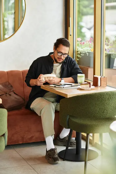 A young man with a beard sits in a charming cafe, focused on work while savoring matcha. — Stock Photo