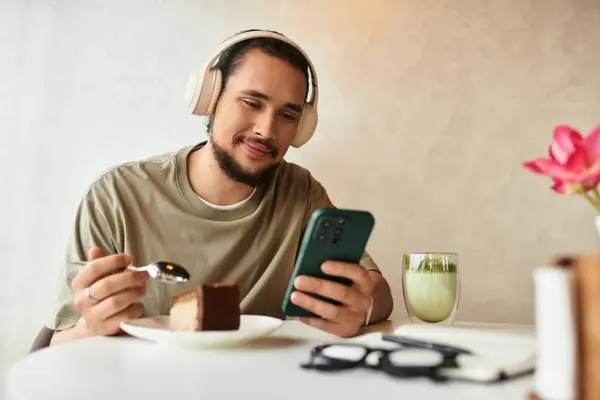 Young man with a beard indulges in cake and matcha while using smartphone at a cafe. — Stock Photo