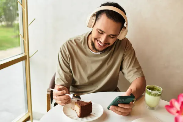 A young man with a beard enjoys matcha and cake while working on his phone at a cafe. — стоковое фото