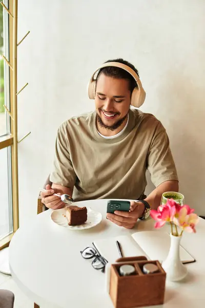 Joven barbudo sonríe mientras usa el teléfono inteligente en un acogedor café, saboreando pastel y matcha. - foto de stock