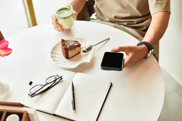 A young man sits at a table, sipping matcha and enjoying cake while working on phone. — Stock Photo