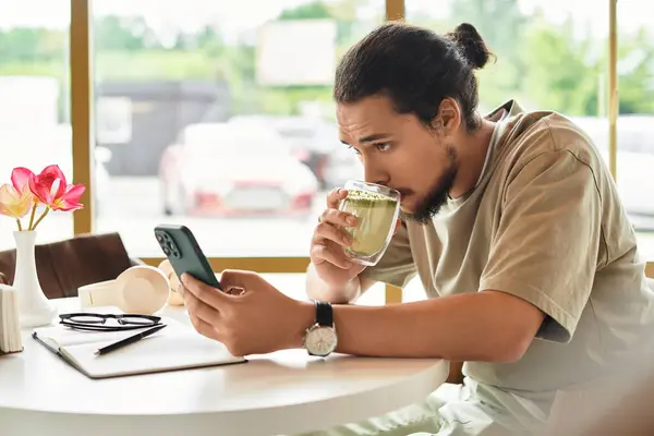 A young man with a beard focuses on his phone, sipping matcha at a cafe. — стоковое фото