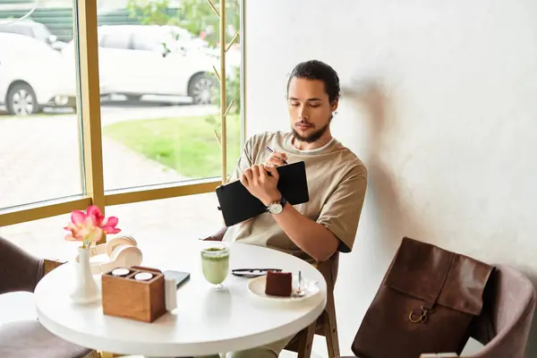 Young man with a beard focused on writing in a notebook while sipping matcha and enjoying dessert. — Stock Photo