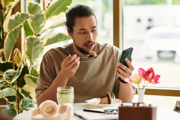 A young man with a beard takes a bite of cake, focused on his phone while enjoying summer in a cafe. — Stock Photo