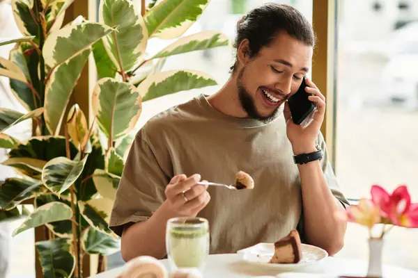 Young man smiles as he takes a call, enjoying cake and matcha in a bright cafe. — Stock Photo