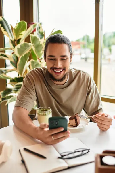 A young man with a beard smiles while checking his phone in a cafe, enjoying matcha and cake. — Stock Photo