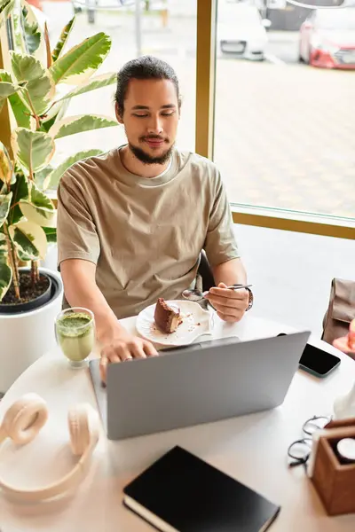 A young man with a beard sits in a cafe, focused on his laptop, enjoying matcha and cake. — Stock Photo