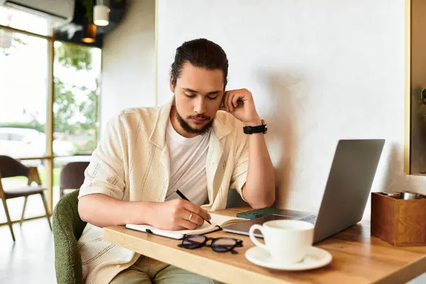 Un joven enfocado con barba escribe notas mientras trabaja remotamente en una mesa de café. — Stock Photo