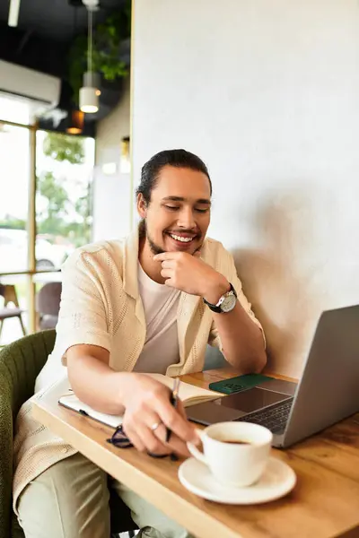 A young man with a beard smiles while working on his laptop at a cafe, sipping coffee. — Stock Photo