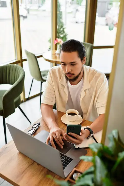Focused young man works remotely at a cafe, sipping coffee and using his laptop and phone. — Stock Photo