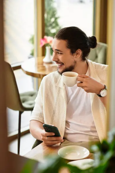 Freelancer with a beard smiles while sipping coffee and using his phone in a cozy cafe. — Stock Photo