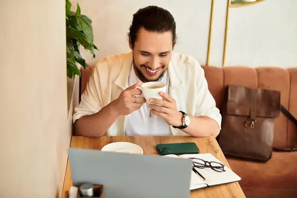 A young man with a beard smiles as he sips coffee while focused on his work in a sunny cafe. — Stock Photo
