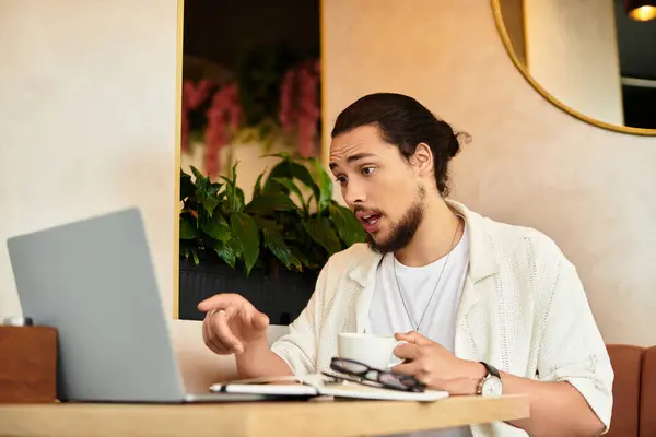 Young man with a beard focused on his laptop, sipping coffee in a lively cafe during summer. — Stock Photo